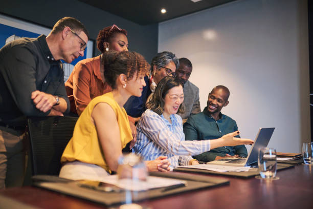 Diverse group of professionals attentively gathered around a table, focused on a laptop screen, showcasing teamwork and engagement in a modern office setting.