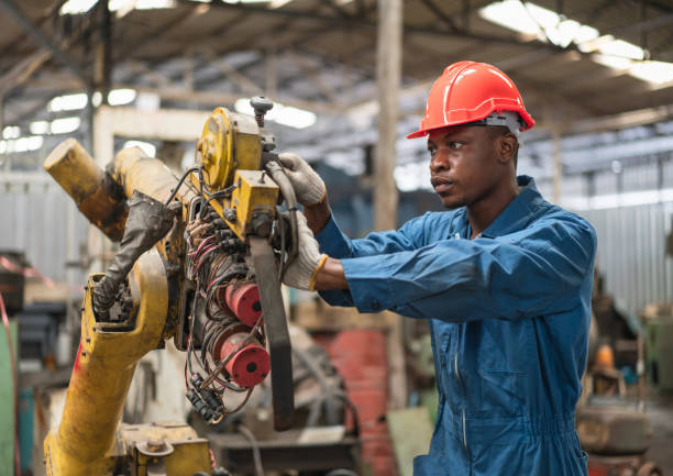 African American male engineer in red safety vest and helmet checking and repairing old robot arm in factory Industrial