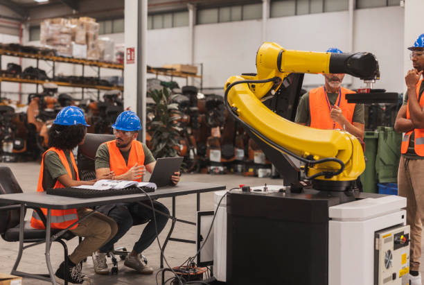 Multiracial team of industrial engineers programming a robotic arm in a large modern factory, discussing and analyzing data on a laptop while another engineer supervises the operation