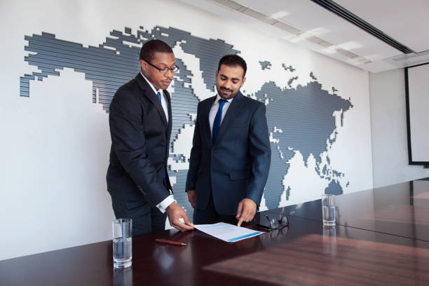 Pair of men team of well dressed suit and tie businessmen partnership executive people in a row at wood conference meeting table talking pointing discussing negotiation planning strategy plans with world map globe earth on wall modern contemporary mural with contracts documents paperwork pens on table prepared decisions choices challenge the way forward future opportunity merger and acquisition combined strength aspirations fine print multi-ethnic Middle Eastern Black